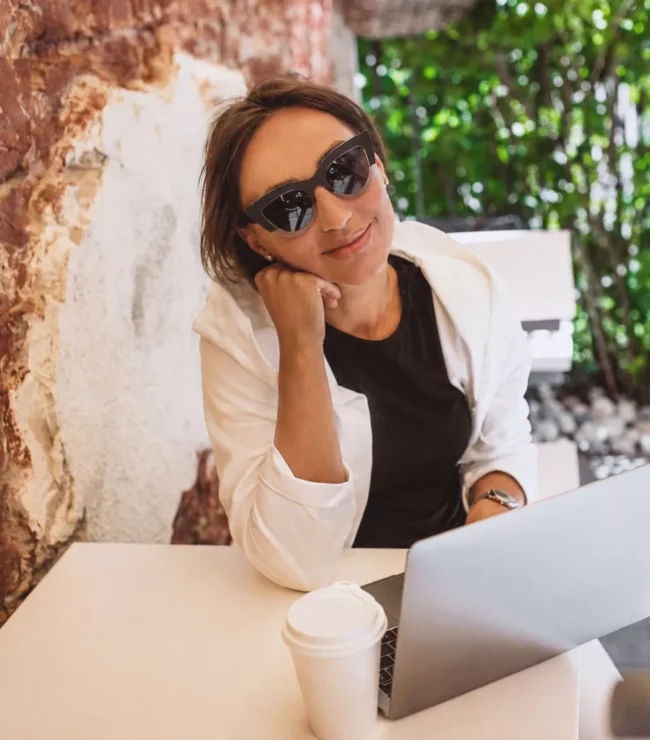 Femme de 40 ans en reconversion professionnelle travaillant sur son ordinateur portable en terrasse, dans une ambiance lumineuse et inspirante. Cette maman active profite d'un moment de calme pour se former, préparer un nouveau projet ou développer son activité, illustrant l'équilibre entre carrière, liberté et slow living.
