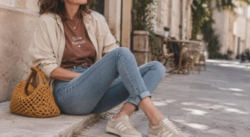Femme de 40 ans au style casual chic assise dans une rue française lumineuse, portant un jean slim bleu clair avec ourlets, un tee-shirt marron « Slow maman », des baskets beige type Gazelle et un sac en maille jaune moutarde.