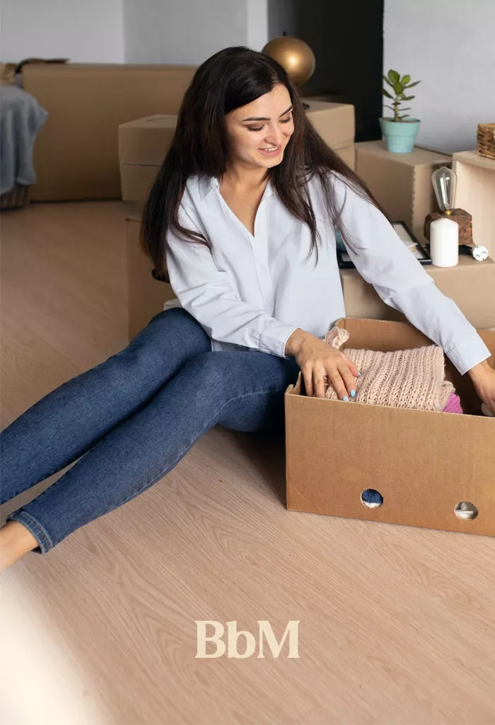 maison organisation rangement mere de famille Femme assise au sol en train de trier et ranger ses vêtements dans une boîte en carton lors d'une séance de désencombrement à la maison, illustration de la règle « 1 entrée = 1 sortie » pour une maison organisée et sereine.