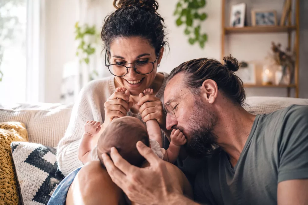 couple apres arrivee bebe jeunes parents complicite slow maman Jeunes parents d'une trentaine d'années partageant un moment de complicité avec leur nouveau-né dans un salon chaleureux, illustration du couple après l'arrivée de bébé selon la philosophie Slow Maman.