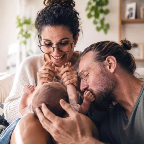 couple apres arrivee bebe jeunes parents complicite slow maman Jeunes parents d'une trentaine d'années partageant un moment de complicité avec leur nouveau-né dans un salon chaleureux, illustration du couple après l'arrivée de bébé selon la philosophie Slow Maman.