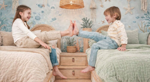 ALT (riche SEO) Deux enfants assis face à face sur leurs lits jumeaux avec housses de couette beige et vert clair, dans une chambre bohème inspirée du monde sous-marin, illustrant la complicité entre frère et sœur dans une chambre partagée.