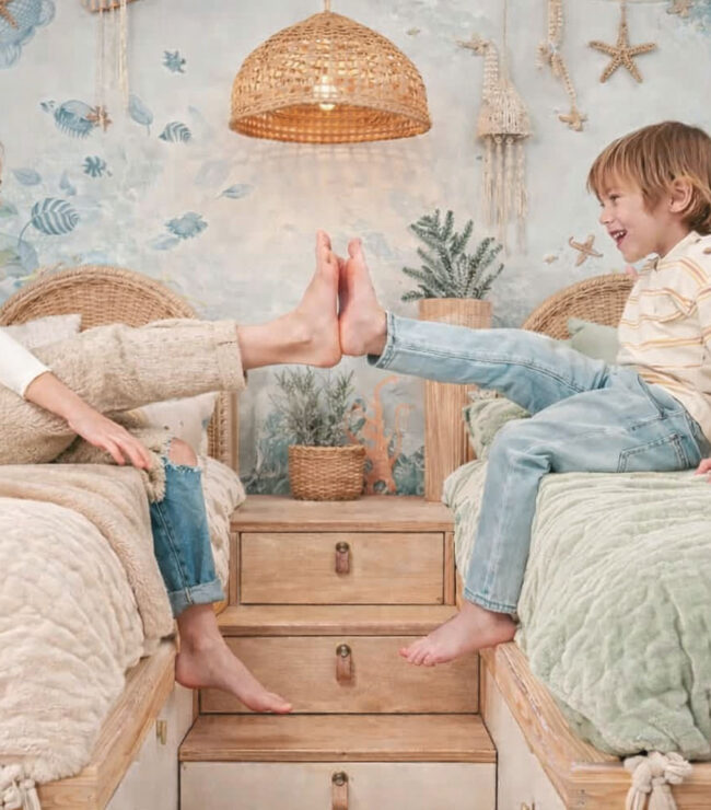 ALT (riche SEO) Deux enfants assis face à face sur leurs lits jumeaux avec housses de couette beige et vert clair, dans une chambre bohème inspirée du monde sous-marin, illustrant la complicité entre frère et sœur dans une chambre partagée.