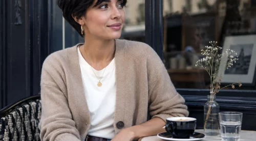 Femme avec une coupe pixie et une frange rideau, look chic et naturel, prenant un café en terrasse dans une ambiance urbaine élégante.
