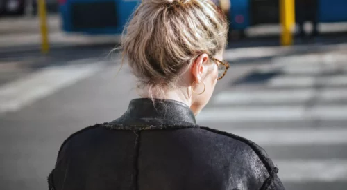 Maman de dos traversant la ville, cheveux blonds attachés en chignon et veste en cuir noir, symbole de la femme moderne entre vie de famille et vie professionnelle.