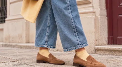 Jean large 7/8e bleu porté avec des mocassins camel en suédine et des chaussettes jaune beurre, devant une façade crème et bordeaux.