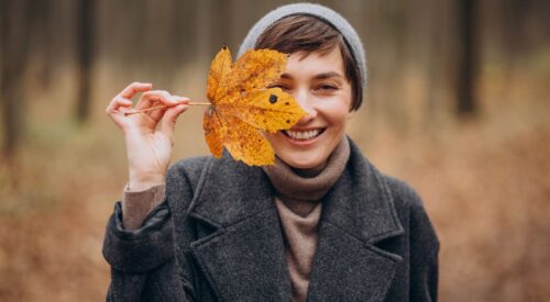 Femme souriante en automne tenant une feuille devant son visage, symbole de bonne humeur, de simplicité et de slow life en pleine nature.