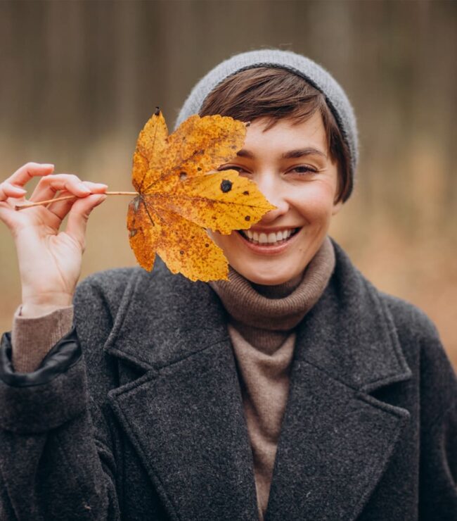 Femme souriante en automne tenant une feuille devant son visage, symbole de bonne humeur, de simplicité et de slow life en pleine nature.