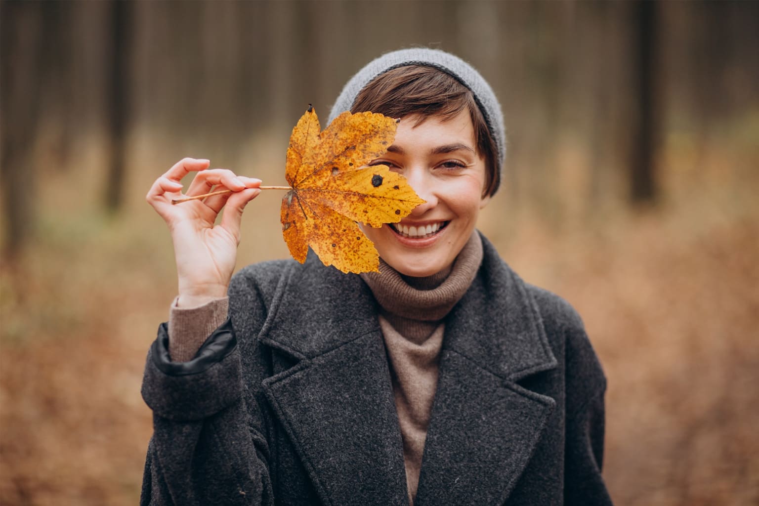 Femme souriante en automne tenant une feuille devant son visage, symbole de bonne humeur, de simplicité et de slow life en pleine nature.