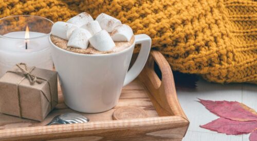 Tasse de chocolat chaud aux marshmallows sur un plateau en bois avec un plaid jaune moutarde, ambiance automne cocooning
