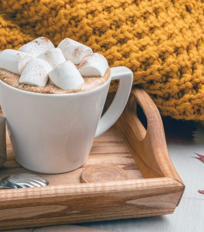 Tasse de chocolat chaud aux marshmallows sur un plateau en bois avec un plaid jaune moutarde, ambiance automne cocooning