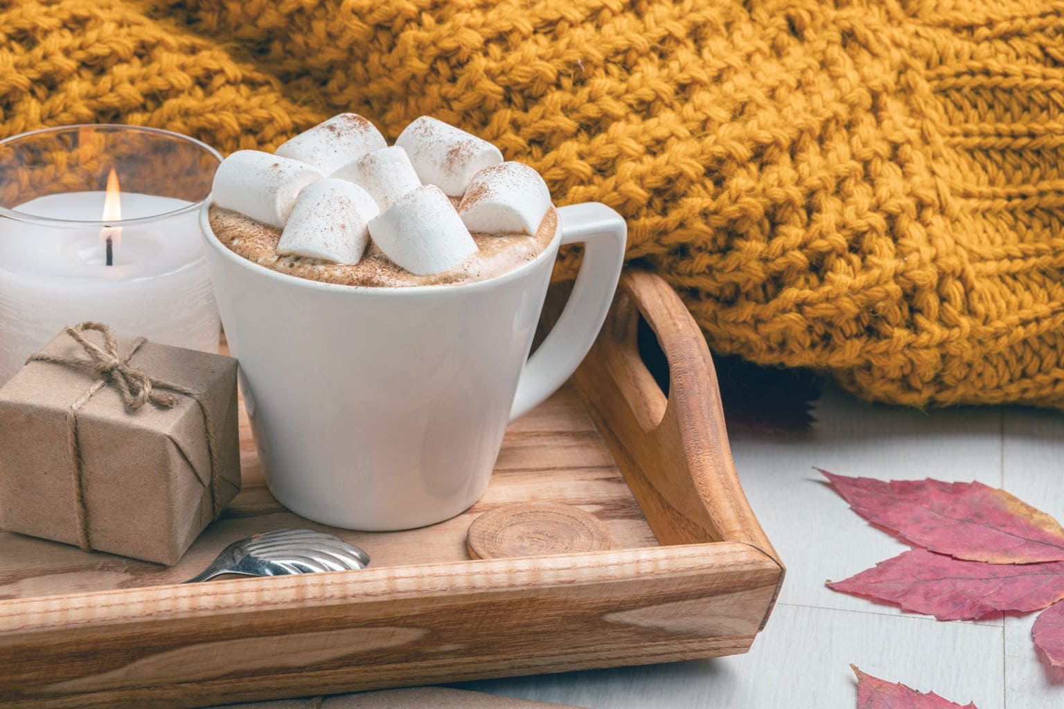 Tasse de chocolat chaud aux marshmallows sur un plateau en bois avec un plaid jaune moutarde, ambiance automne cocooning