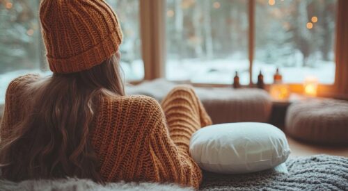 Femme en mode cocooning à la maison, emmitouflée dans un pull et un bonnet face à la neige