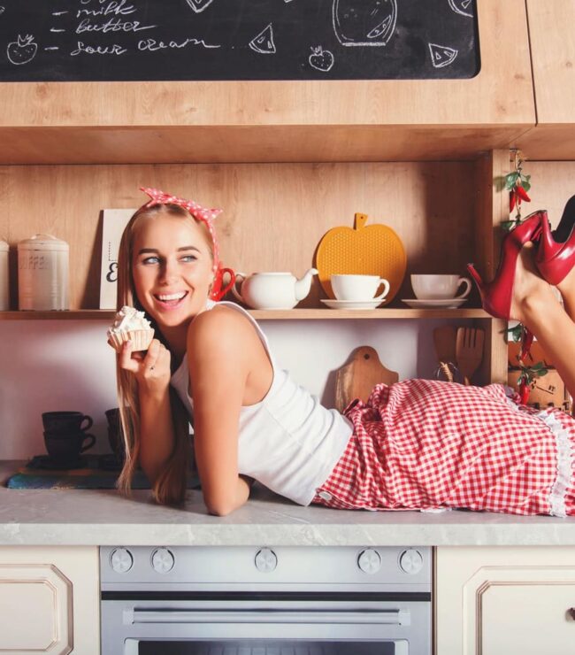 Femme au look rétro allongée sur un plan de travail, souriante avec un cupcake dans une cuisine vintage