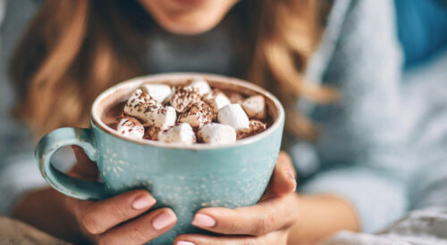 Femme souriante tenant une grande tasse de chocolat chaud garnie de chamallows, symbole d’un moment cocooning et chaleureux en hiver.