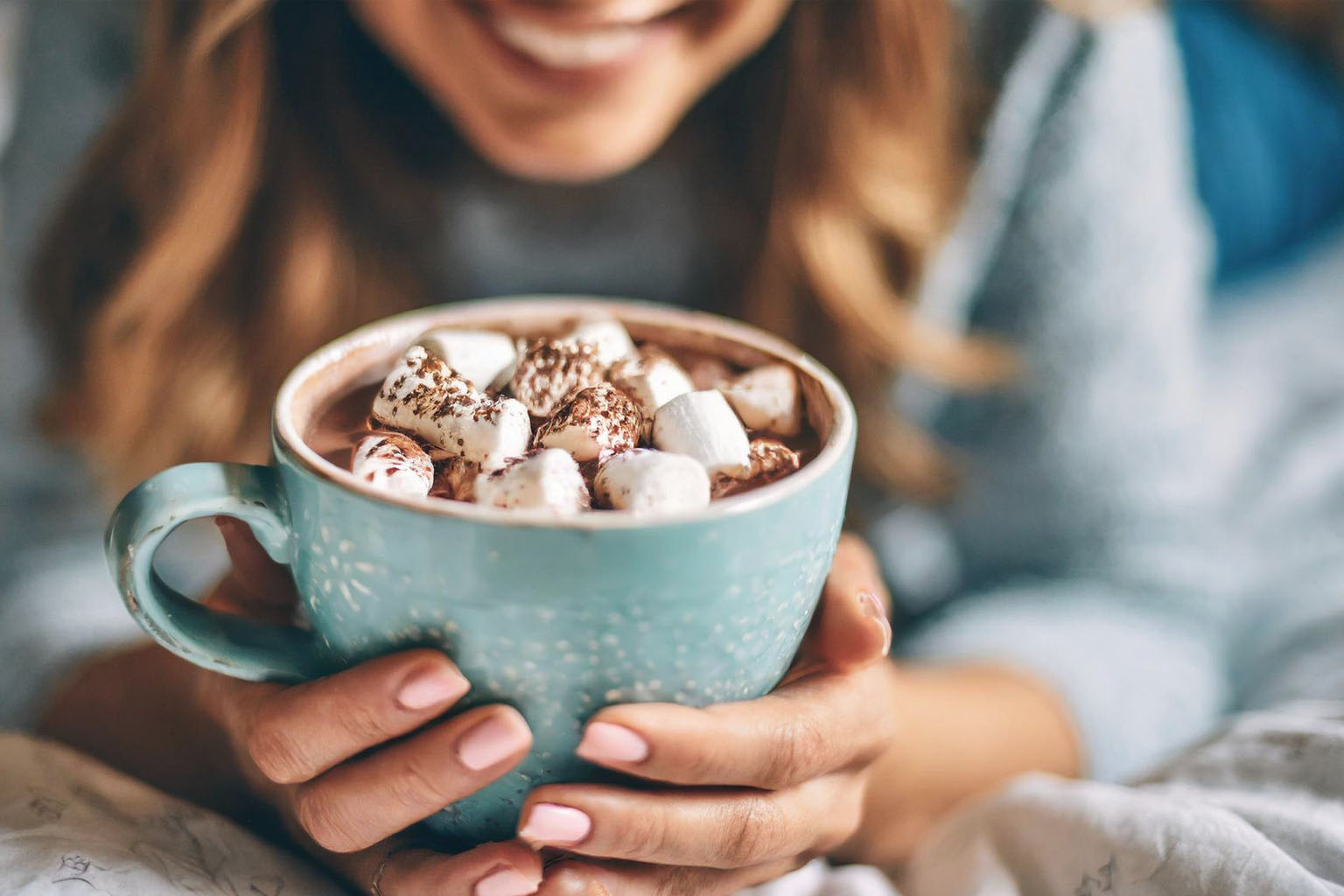 Femme souriante tenant une grande tasse de chocolat chaud garnie de chamallows, symbole d’un moment cocooning et chaleureux en hiver.