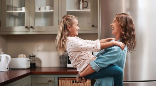 Maman souriante qui porte sa fille dans la cuisine, moment de complicité familiale et d’équilibre entre amour et bien-être.