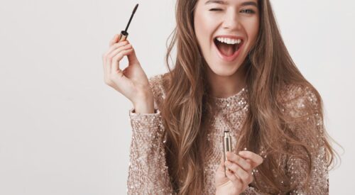 Femme souriante en tenue à paillettes appliquant du mascara, maquillage glamour pour le réveillon du 31 décembre.
