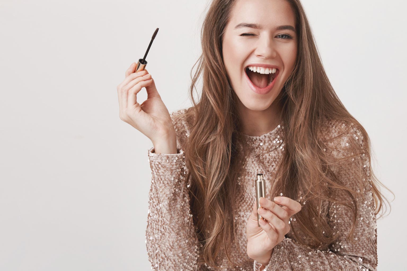 Femme souriante en tenue à paillettes appliquant du mascara, maquillage glamour pour le réveillon du 31 décembre.