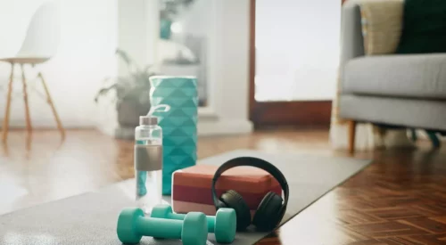 Tapis de yoga avec haltères, gourde et casque audio dans un salon lumineux, illustrant une routine sportive simple et accessible à pratiquer à la maison quand on est maman.