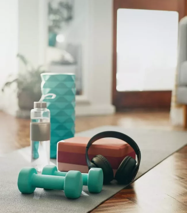 Tapis de yoga avec haltères, gourde et casque audio dans un salon lumineux, illustrant une routine sportive simple et accessible à pratiquer à la maison quand on est maman.