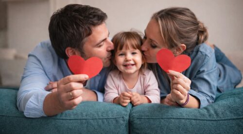 Famille souriante célébrant la Saint-Valentin à la maison, parents et enfant appuyés sur le dossier d’un canapé, partageant un moment complice