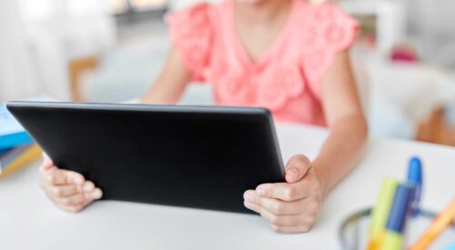 Enfant utilisant une tablette numérique à l’école, assise à un bureau avec des fournitures scolaires, symbole de la scolarité connectée et des écrans en classe.