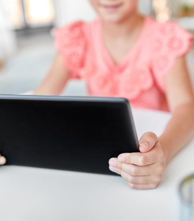 Enfant utilisant une tablette numérique à l’école, assise à un bureau avec des fournitures scolaires, symbole de la scolarité connectée et des écrans en classe.