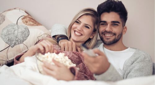 Couple souriant allongé dans un lit regardant la télévision, moment de détente et de complicité dans la chambre parentale