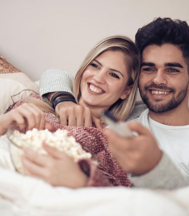 Couple souriant allongé dans un lit regardant la télévision, moment de détente et de complicité dans la chambre parentale