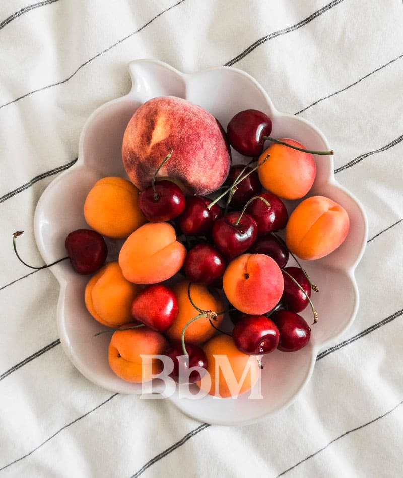 corbeille fruit maison printemps goodvibes bebymaman Corbeille de fruits de saison posée sur une table, avec abricots, cerises et pêche dans une assiette en céramique claire, ambiance printanière.