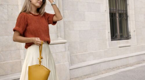 Femme blonde cendrée portant un pull terracotta et une jupe longue écrue, style chic et intemporel, sac moutarde à la main