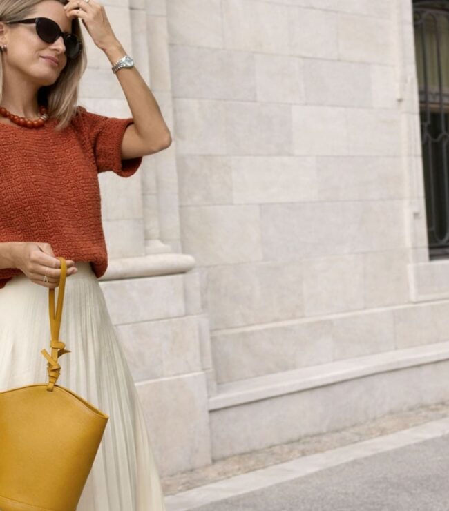 Femme blonde cendrée portant un pull terracotta et une jupe longue écrue, style chic et intemporel, sac moutarde à la main