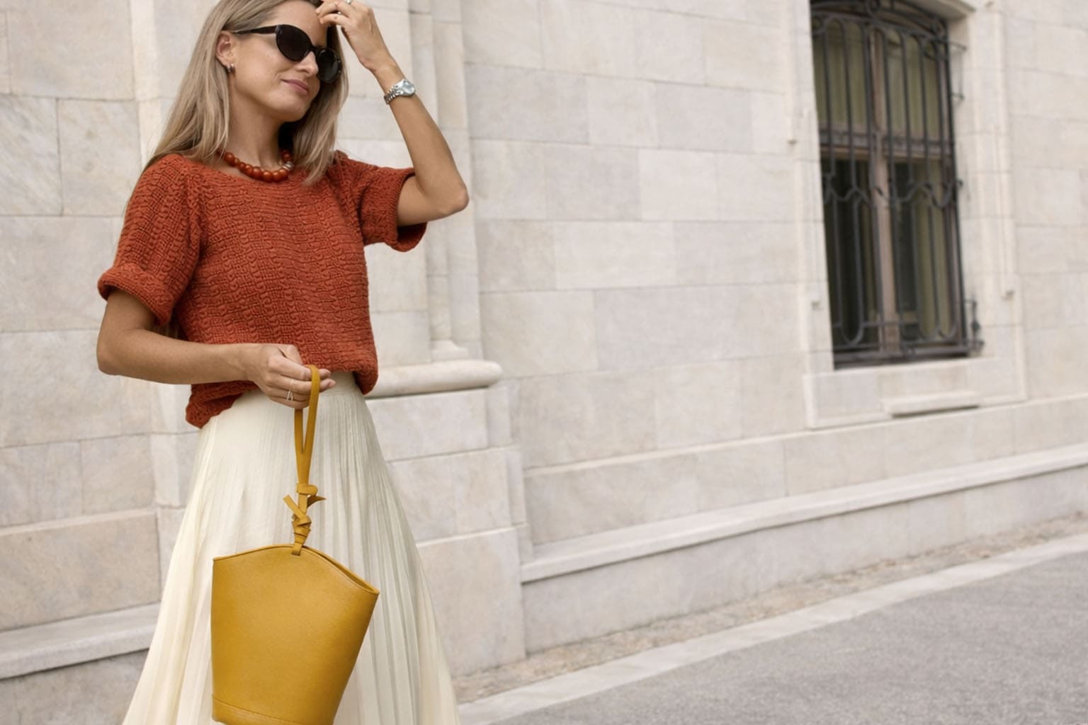 Femme blonde cendrée portant un pull terracotta et une jupe longue écrue, style chic et intemporel, sac moutarde à la main