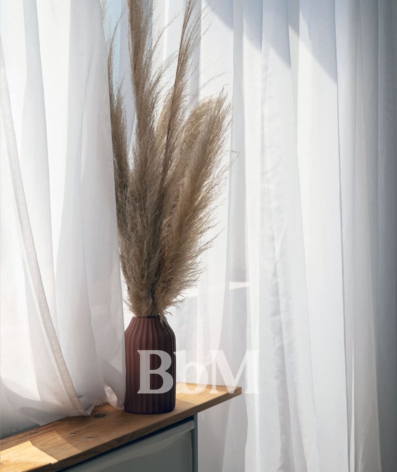 rideau voilage lumiere maison printemps goodvibes bebymaman Voilages blancs légers laissant passer la lumière naturelle du printemps dans une pièce, avec un vase en céramique et des herbes séchées posé sur un rebord de fenêtre.