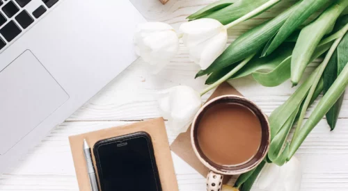 Espace de travail slow working avec ordinateur portable, café, carnet, smartphone et tulipes blanches, illustrant un mode de travail plus serein pour une maman quadra en quête d'équilibre entre vie professionnelle et vie personnelle.