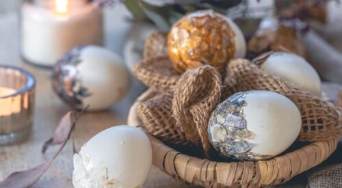 Décoration de Pâques chic avec œufs décoratifs dorés et nacrés dans un panier en osier, bougies allumées et matières naturelles sur une table rustique.
