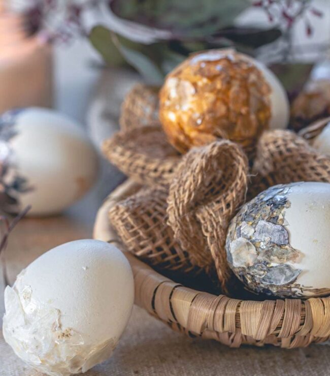 Décoration de Pâques chic avec œufs décoratifs dorés et nacrés dans un panier en osier, bougies allumées et matières naturelles sur une table rustique.