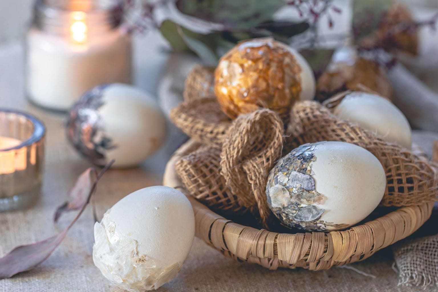 Décoration de Pâques chic avec œufs décoratifs dorés et nacrés dans un panier en osier, bougies allumées et matières naturelles sur une table rustique.