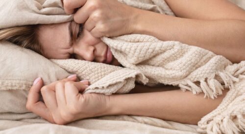 Femme allongée dans des draps beige et un plaid bohème crème, se cachant partiellement le visage dans la couette, ambiance cocooning et glamour, illustrant la fatigue pendant les règles.