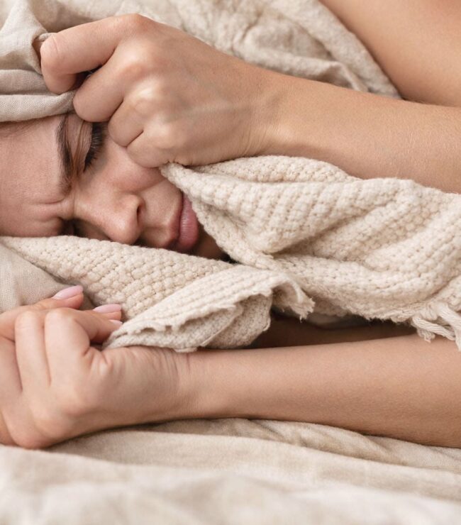 Femme allongée dans des draps beige et un plaid bohème crème, se cachant partiellement le visage dans la couette, ambiance cocooning et glamour, illustrant la fatigue pendant les règles.