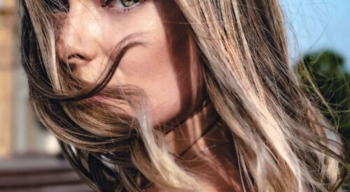 Femme aux cheveux châtains lumineux au vent, regard vert intense, beauté naturelle printemps-été 2026