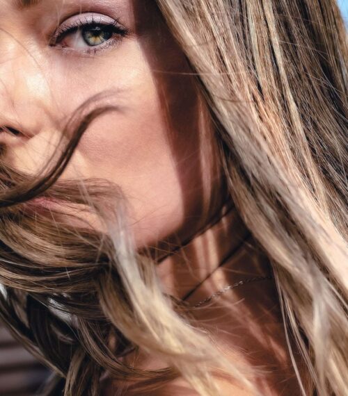 Femme aux cheveux châtains lumineux au vent, regard vert intense, beauté naturelle printemps-été 2026