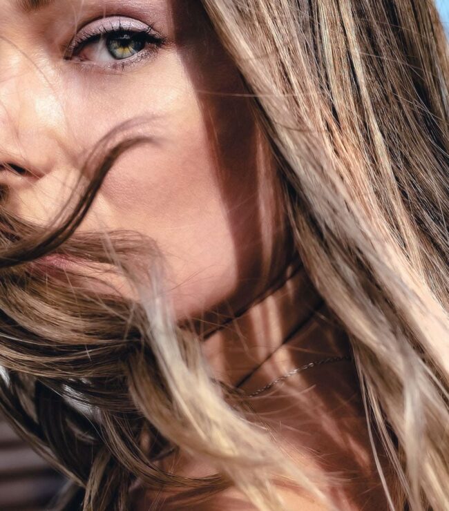 Femme aux cheveux châtains lumineux au vent, regard vert intense, beauté naturelle printemps-été 2026