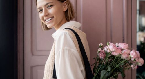 Maman en look casual chic avec blouse blanche fluide et top en maille beige sans manches, portant un tote bag avec des fleurs