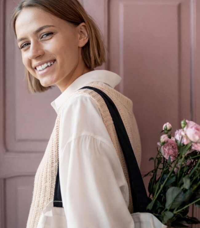 Maman en look casual chic avec blouse blanche fluide et top en maille beige sans manches, portant un tote bag avec des fleurs