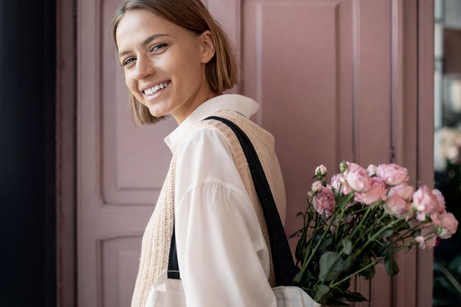 Maman en look casual chic avec blouse blanche fluide et top en maille beige sans manches, portant un tote bag avec des fleurs