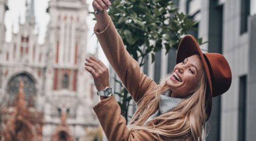 Femme souriante prenant un selfie en ville avec un chapeau, illustration d’une maman hybride moderne qui assume ses différentes vies entre maternité, travail et passions.