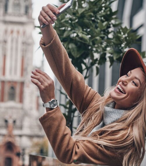 Femme souriante prenant un selfie en ville avec un chapeau, illustration d’une maman hybride moderne qui assume ses différentes vies entre maternité, travail et passions.