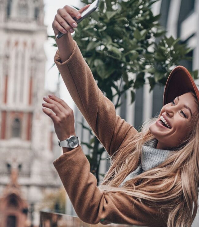 Femme souriante prenant un selfie en ville avec un chapeau, illustration d’une maman hybride moderne qui assume ses différentes vies entre maternité, travail et passions.
